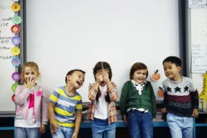 A diverse group of young children laughing and playing together in front of a whiteboard in a bright classroom setting.