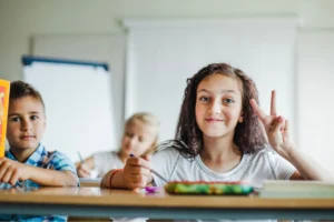 A smiling young girl sitting at a classroom desk, holding a pencil and making a peace sign with her hand, with other children blurred in the background.