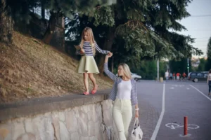 A mother and daughter walking together hand-in-hand on a sidewalk in Madrid; the young girl walks along a low stone wall while holding a tablet.
