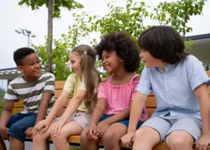 A diverse group of four happy children sitting on an outdoor wooden bench, laughing and talking together in a sunny park setting.