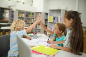 Three happy children in a bright library setting, with two of them giving each other a high five over their desks while smiling.
