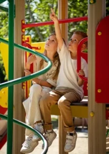 Two young children sitting on a colorful playground structure, looking up and pointing with curiosity at something in the distance during a sunny day.