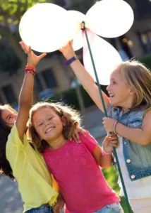 Three happy children outdoors holding white balloons and celebrating with joy in a sunny Madrid park.