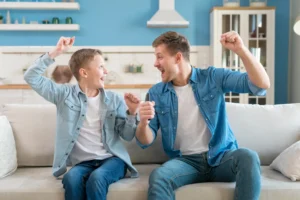 A father and his son sitting on a sofa and celebrating enthusiastically, capturing a moment of emotional support and connection at home.