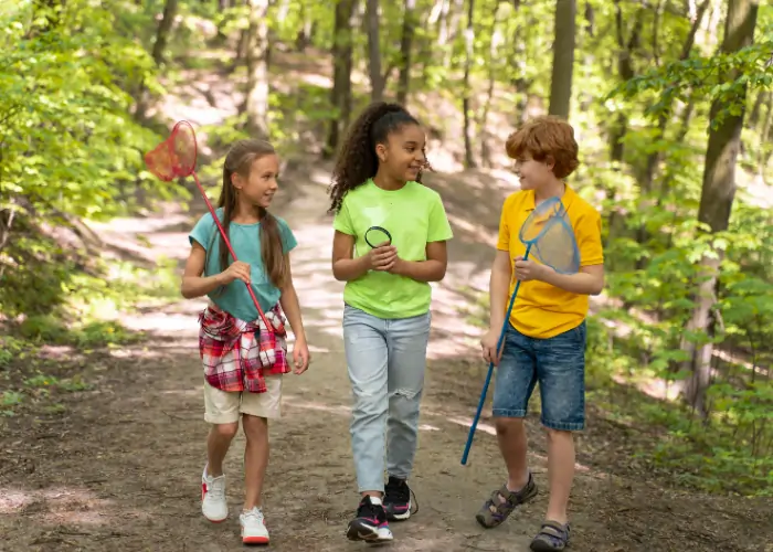 Three children walking together along a wooded trail, carrying colorful butterfly nets and engaging in a friendly conversation.