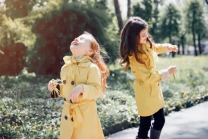 Two young girls in yellow raincoats laughing and playing outdoors in a green park, holding dandelions and enjoying a moment of carefree friendship.