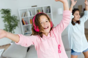 Two young girls laughing and dancing with pink and white headphones on, illustrating how music and rhythm help children internalize a new language through play and movement.