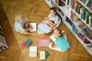 hree children sitting on a wooden floor in a Madrid library surrounded by books, smiling and learning Spanish together.