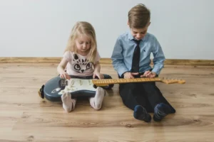 Two young children sitting on a wooden floor playing together with a guitar in a low-pressure learning environment.