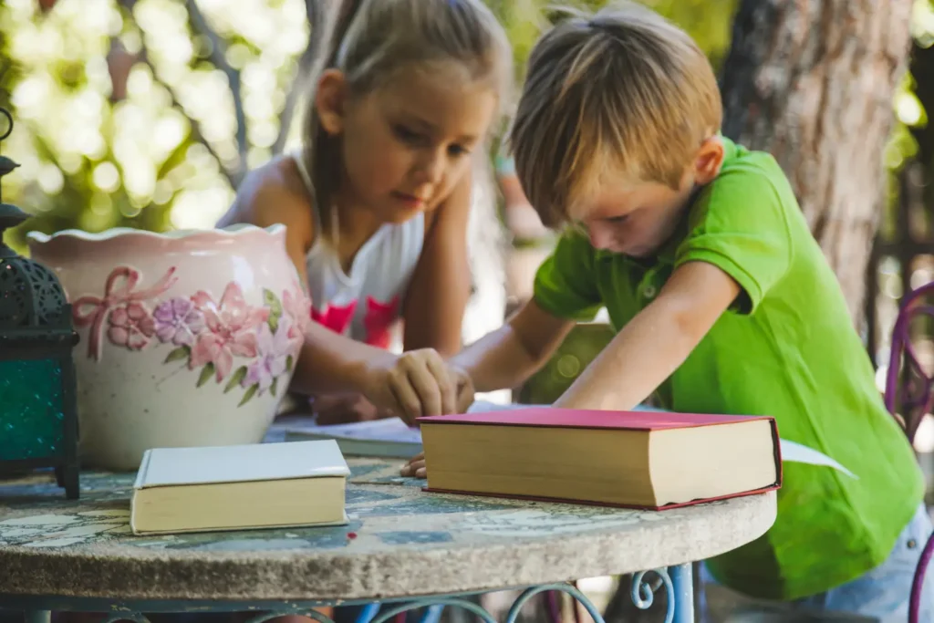 A young girl and a young boy in a green shirt leaning over a mosaic stone table outside, working intently on an interactive project with books nearby.
