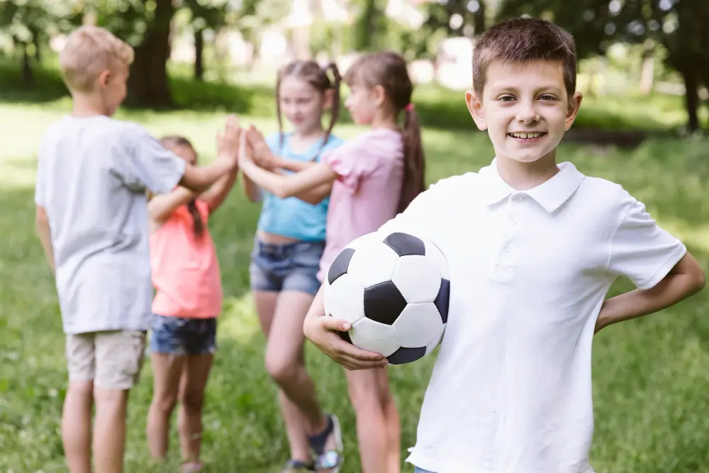 A smiling young boy in a white polo shirt holding a classic black and white football tucked under his arm, with a blurred group of children high-fiving in a sunny park background.