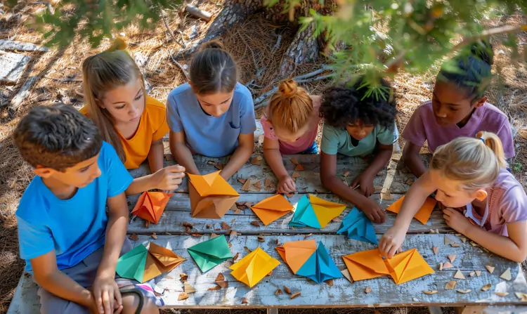 A top-down view of a small, diverse group of seven children sitting around a rustic wooden table outdoors in a wooded park, folding colorful paper origami shapes together.