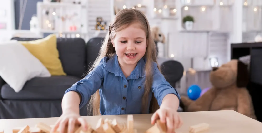 A young girl with long hair smiling as she plays with scattered wooden blocks on a table in a brightly lit, cozy living room.