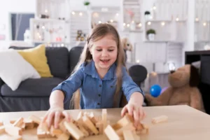A young girl with long hair smiling as she plays with scattered wooden blocks on a table in a brightly lit, cozy living room.