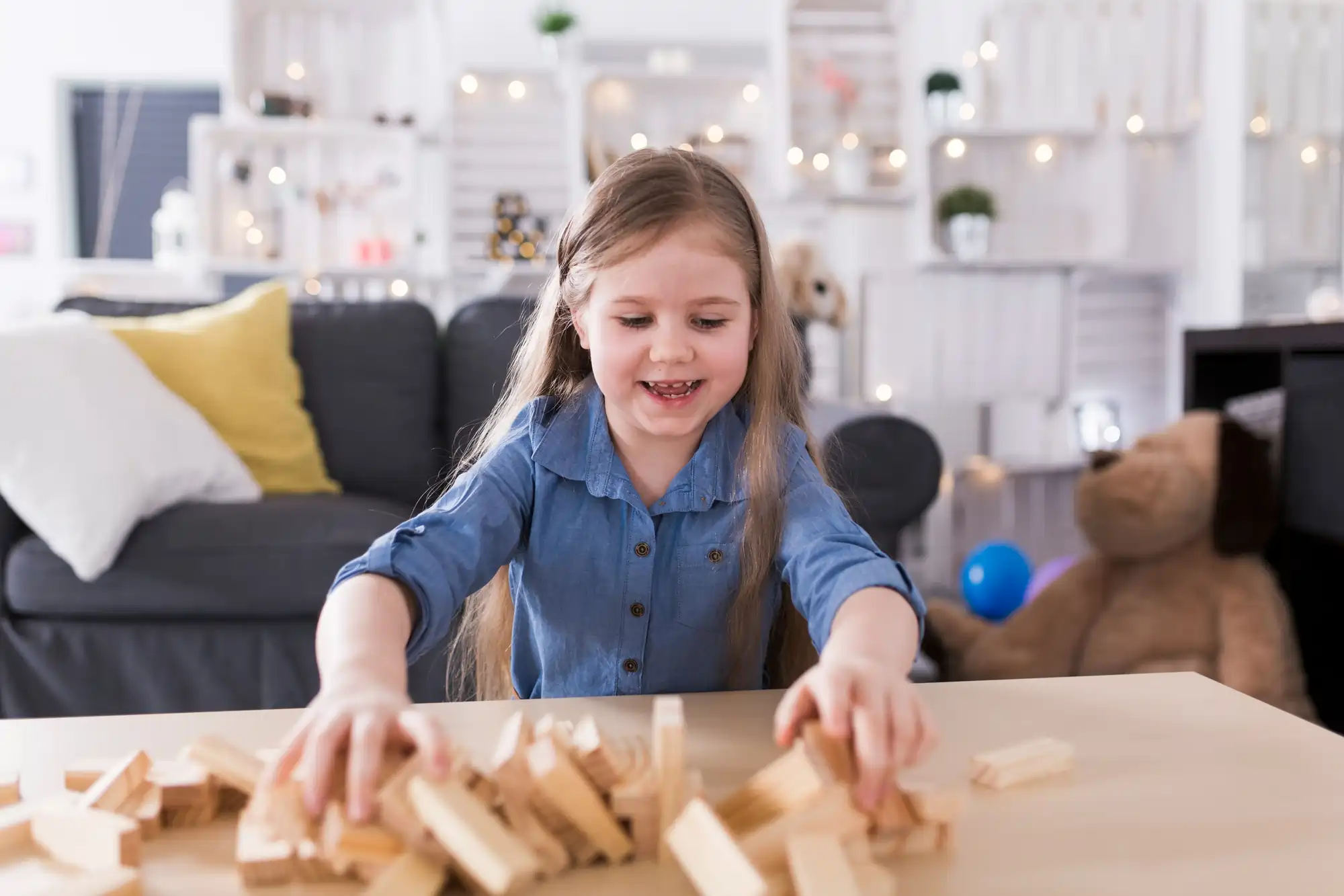 A young girl with long hair smiling as she plays with scattered wooden blocks on a table in a brightly lit, cozy living room.
