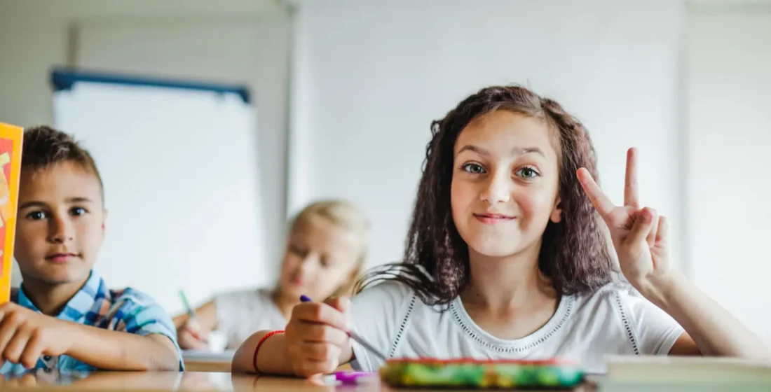 A smiling young girl sitting at a classroom desk, holding a pencil and making a peace sign with her hand, with other children blurred in the background.