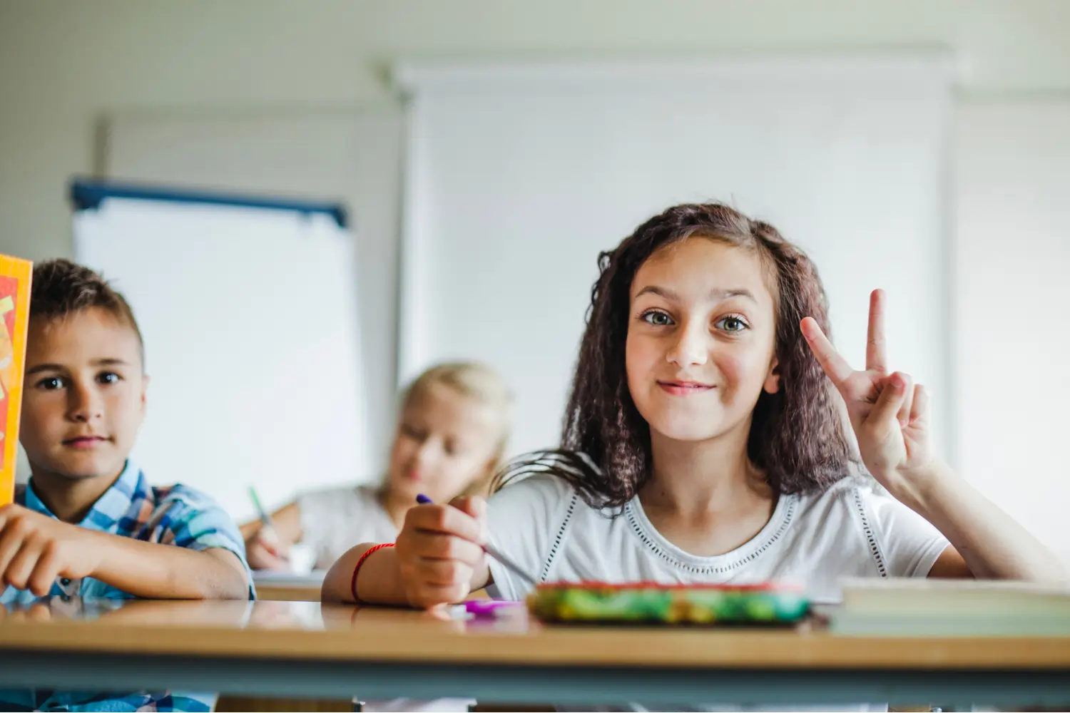 A smiling young girl sitting at a classroom desk, holding a pencil and making a peace sign with her hand, with other children blurred in the background.