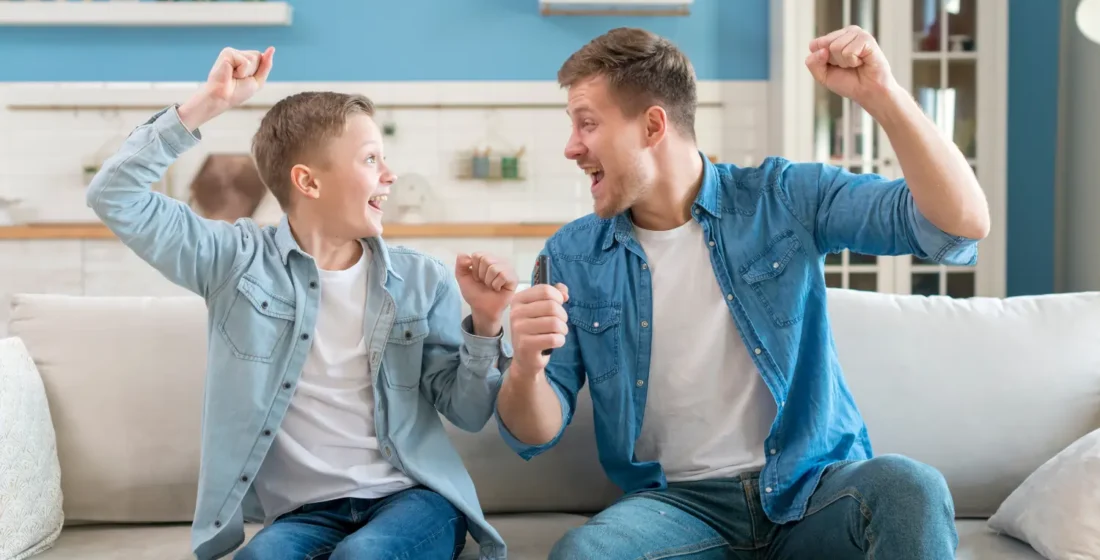 A father and his son sitting on a sofa and celebrating enthusiastically, capturing a moment of emotional support and connection at home.