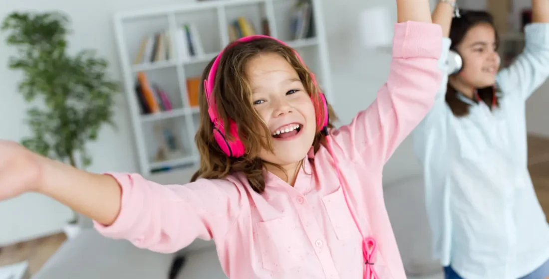 Two young girls laughing and dancing with pink and white headphones on, illustrating how music and rhythm help children internalize a new language through play and movement.