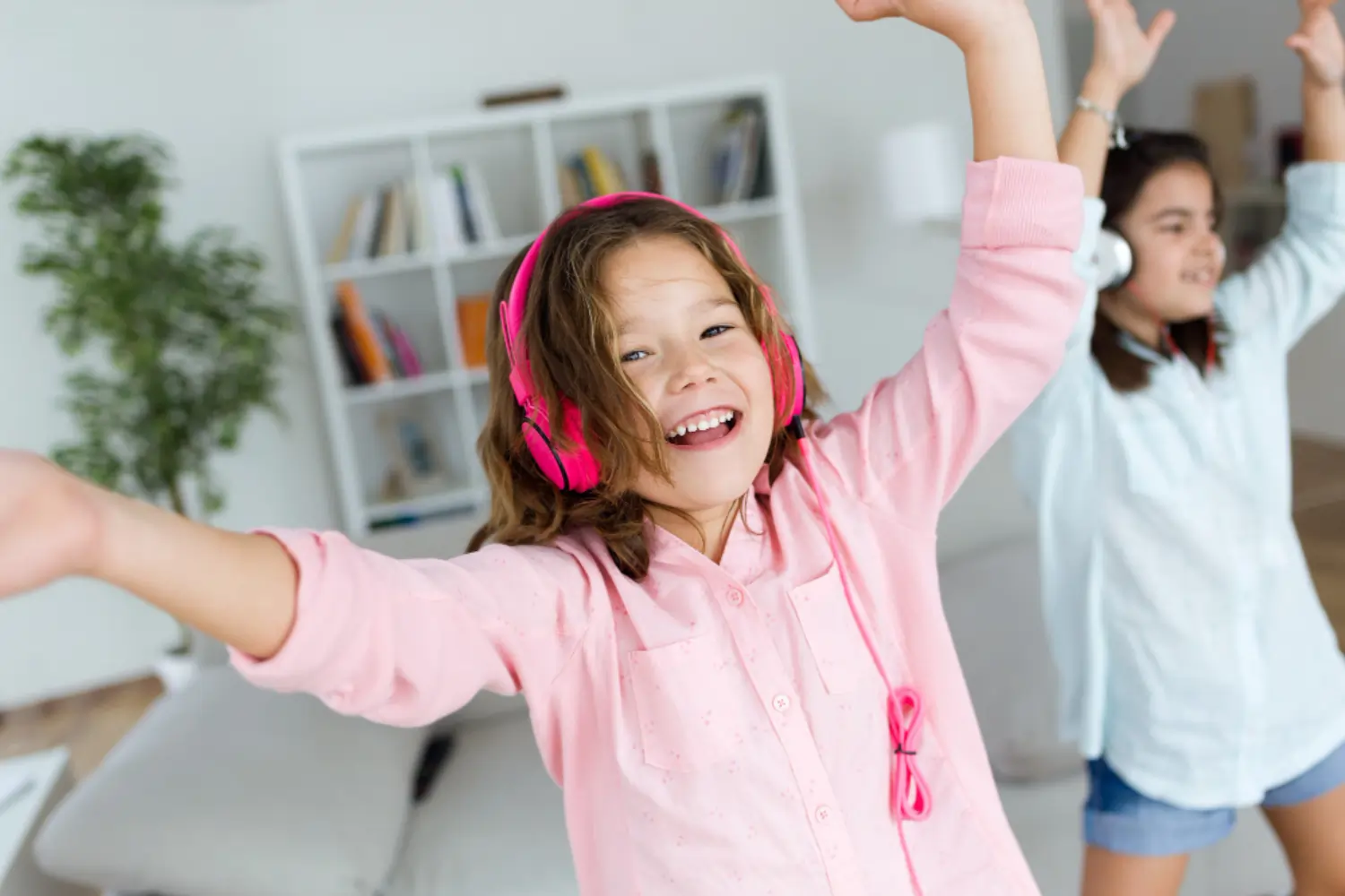Two young girls laughing and dancing with pink and white headphones on, illustrating how music and rhythm help children internalize a new language through play and movement.