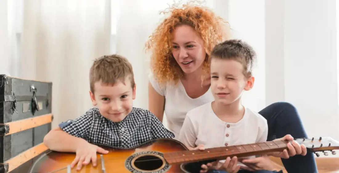 Two young children sitting on a wooden floor playing together with a guitar in a low-pressure learning environment.