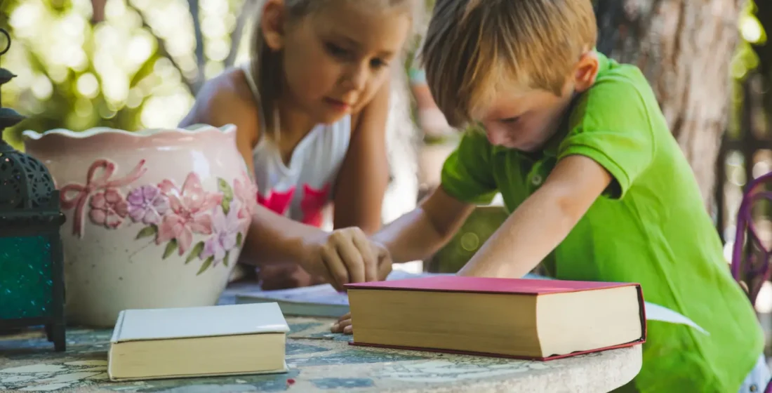 A young girl and a young boy in a green shirt leaning over a mosaic stone table outside, working intently on an interactive project with books nearby.