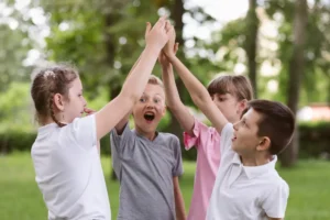 Four happy, excited children laughing and high-fiving together outdoors in a sunny, green tree-lined park.