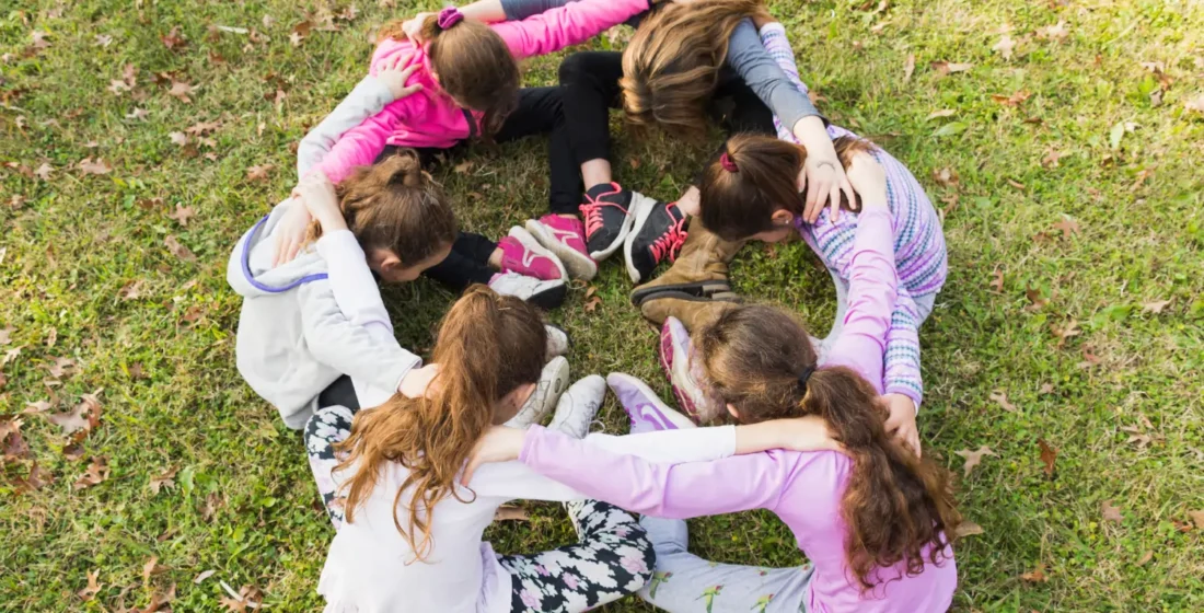 Un grupo pequeño de niños sentados en círculo sobre el césped de un parque, abrazados por los hombros en una piña muy unida.