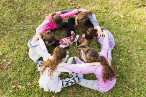 Un grupo pequeño de niños sentados en círculo sobre el césped de un parque, abrazados por los hombros en una piña muy unida.