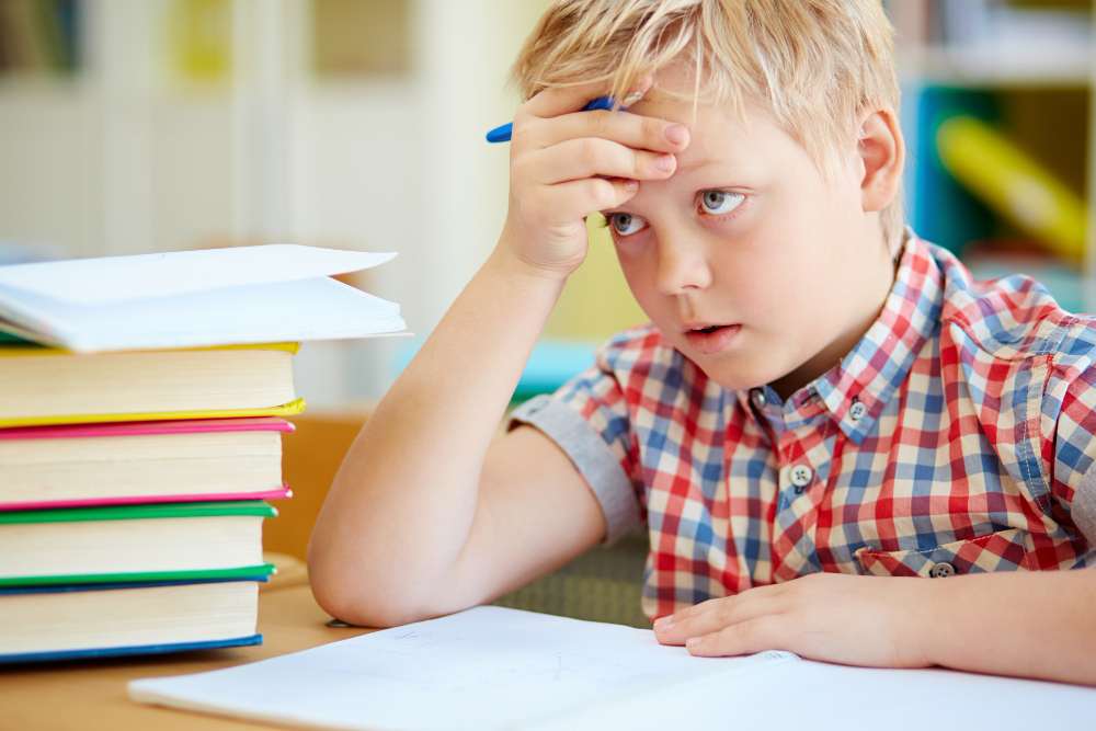 A young boy resting his hand on his head with a confused, overwhelmed expression sitting in front of a stack of school textbooks.