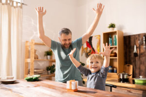 A young boy and his father joyfully raising their arms in celebration at a wooden kitchen table.