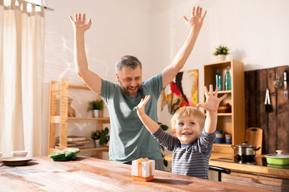 A young boy and his father joyfully raising their arms in celebration at a wooden kitchen table.